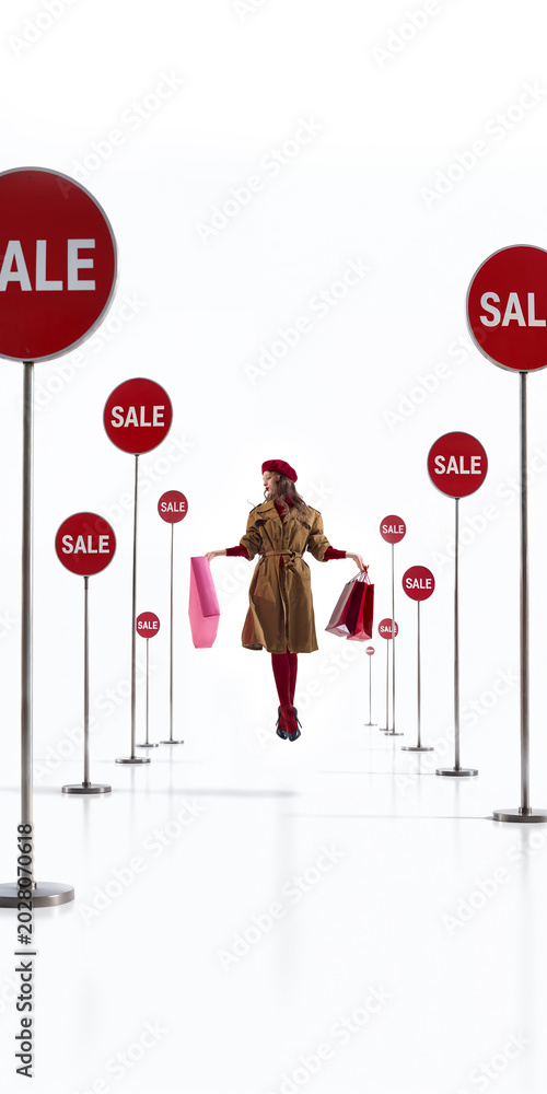 Obraz premium Fashion shopper surrounded by red sale signs holding bags in white studio. Concept of retail overload, discount season, consumer pressure, shopping temptation and promo design.