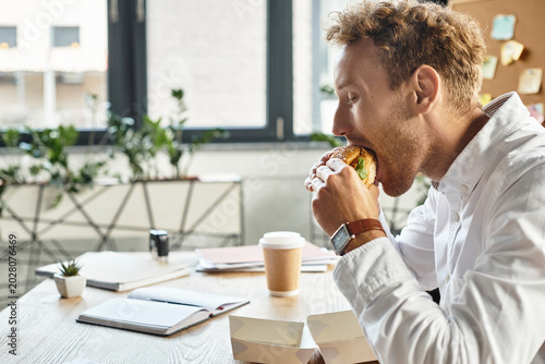 Young man enjoying a delicious burger while working in a cozy modern workspace