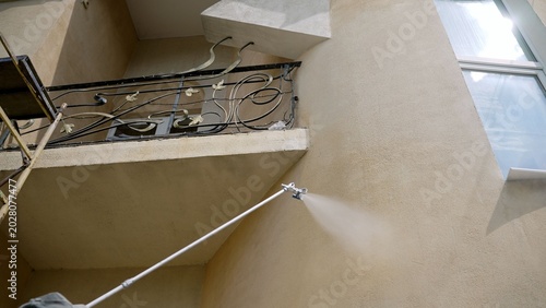 Worker paints building facade with airless spray gun. Professional painter applies beige paint on stucco wall using extension pole. House renovation and exterior improvement process