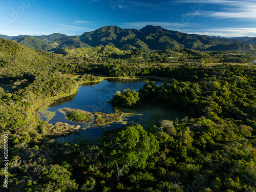 Aerial view showing calm lake water surrounded by lush green tropical trees and vibrant foliage with distant mountains under a blue sky, highlighting an untouched natural ecosystem, Chiriqui, Panama.