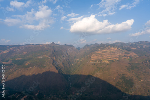 Vast mountain range under bright blue sky. Soft clouds drift above rugged peaks. Deep shadow falls across rolling valleys. Terraced fields dot the slopes below. Natural landscape untouched and serene