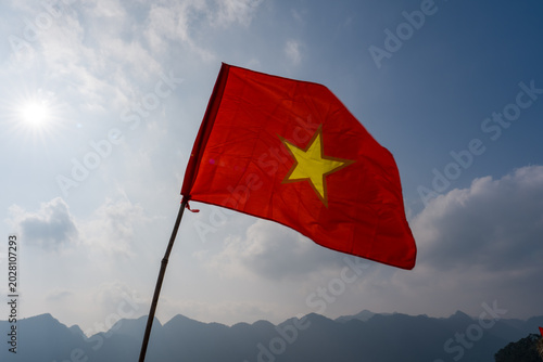 Red flag with yellow star waves in mountain breeze. Bright sun shines behind distant hills. Flagpole stands tall against blue sky. Clouds drift softly in background
