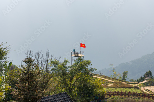 Red flag flies atop a tower in misty mountains. Green trees frame the foreground with soft focus. Misty hills stretch behind the structure in hazy tones. Rustic roofline peeks from beneath the foliage