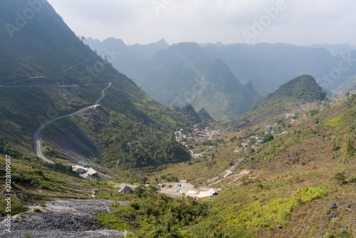 Winding road cuts through lush green mountains. Village nestled in valley below. Rugged peaks rise under soft, cloudy sky. Vast, tranquil landscape invites exploration. No animals or plants visible