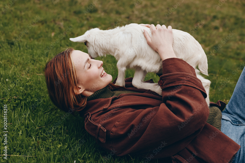 Fototapeta premium A young woman with red hair lies on grass smiling as she holds a white baby goat close to her face in a casual outdoor lifestyle moment.