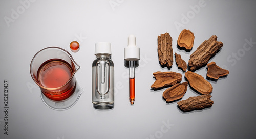 Cinchona bark extract preparation displayed with a laboratory beaker, medicinal oil dropper bottle, and raw wood chips arranged on a clean gray surface