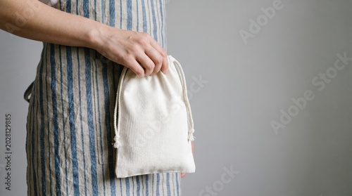 Person in striped apron holding a white fabric drawstring bag. Sustainable zero waste lifestyle and eco-friendly shopping concept, horizontal with copy space.