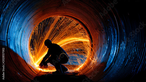 A worker creates sparks while welding inside a large metal pipe in an industrial area at night. The sparks light up the surroundings with bright colors