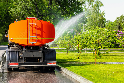 Orange water sprinkler truck watering trees in a park. A special machine waters the lawn in the park. a car sprays water on trees in a park. A watering machine waters trees on a hot day