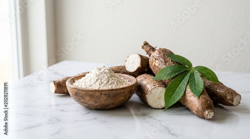 Fresh cassava roots and a wooden bowl filled with starchy white flour on marble table