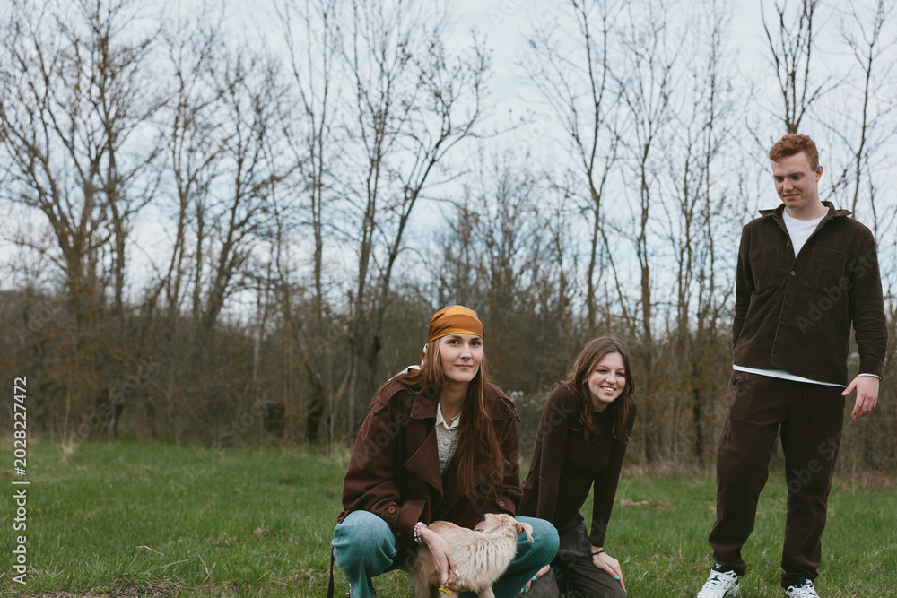 Fototapeta premium Three young adults share a playful moment with a goat kid in a grassy park, casual clothing, natural light, candid lifestyle shot capturing friendship, rural charm, and simple outdoor joy.