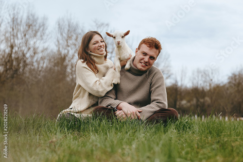 Obraz na plátne Smiling couple sitting on grass in an open field with a playful goat kid between