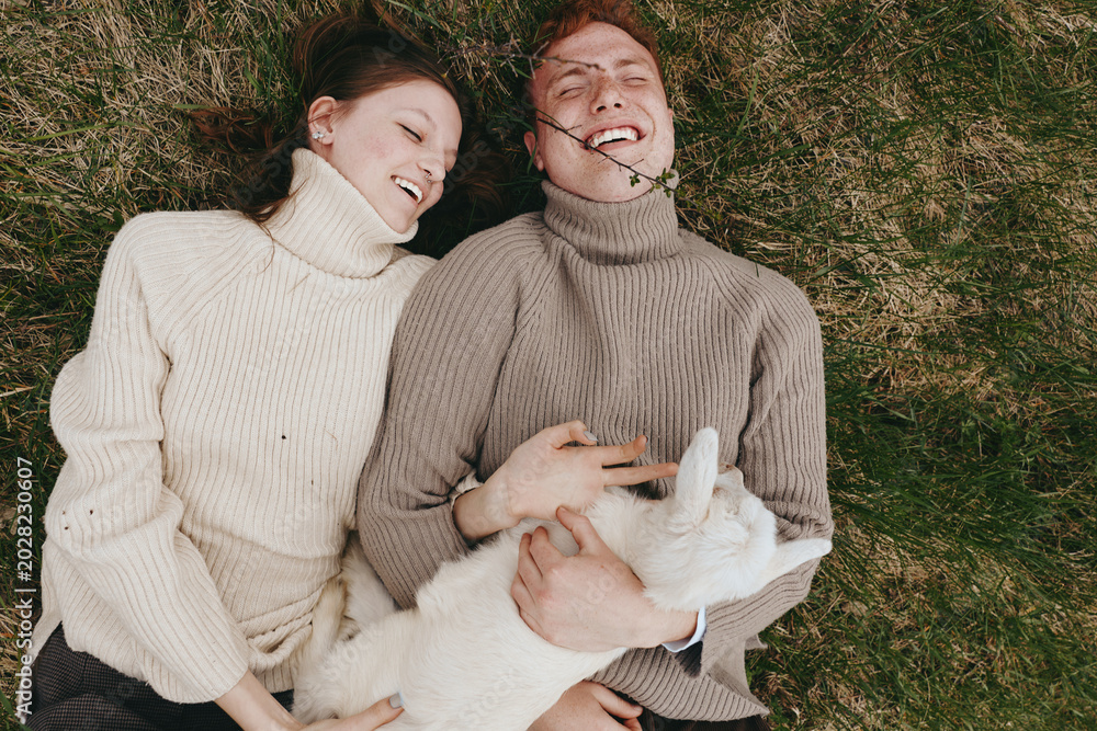 Fototapeta premium Two smiling adults lie on green grass wearing chunky sweaters, enjoying a cozy outdoor moment together with a small animal between them, showcasing lifestyle comfort, warmth, and carefree affection.