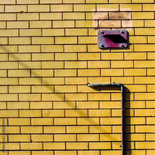 Saturated yellow painted brick wall with teal pipe elbow and rust-coloured mail box and ghost rectangle mark