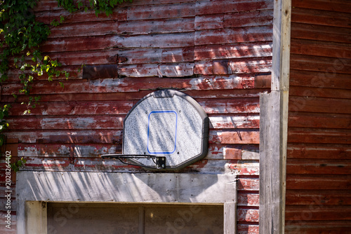 Принти на полотні A worn basketball hoop and backboard are mounted on the weathered red siding of a rural barn, a quietly evocative image of backyard sports and small-town American childhood