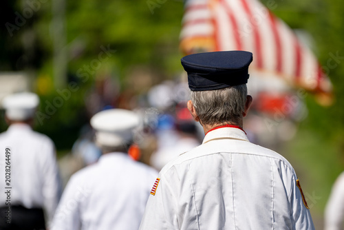 Canvas Print An older man in a white uniform and black cap is seen from behind at a Memorial Day or veteran's parade, American flags and what appears to be a fire truck visible ahead of him in the procession