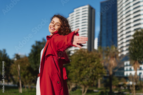 Young woman in a red coat smiles with open arms in an urban park, sunlight highlights her expression as modern buildings rise behind, conveying freedom.