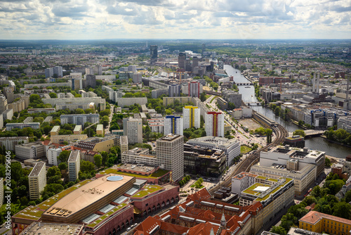 Aerial panoramic view of Berlin, Germany, with urban skyline, river, bridges, residential buildings, green parks and dramatic cloudy sky, showing the cityscape from above.