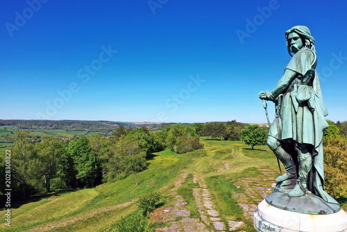 Εκτύπωση καμβά Das monumentale Vercingétorix-Denkmal auf einem steinernen Sockel, eingebettet in eine grüne Hügellandschaft mit Bäumen unter einem strahlend blauen Himmel