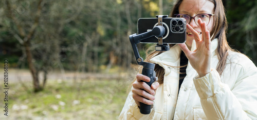 Woman filming with a smartphone gimbal in a forest. Mobile video creation in outdoor natural light. Travel vlogging and social media content concept..