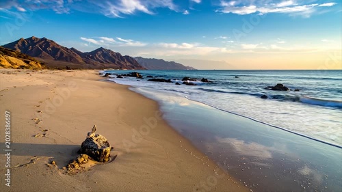 Sandy beach with mountains, calm ocean, and blue sky. Footprints in sand, rocks
