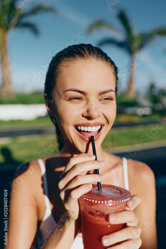 Schilderij op canvas A young woman in a white tank top smiles while sipping a pink smoothie in a sunlit outdoor setting, conveying a casual lifestyle moment with relaxed posture and a vibrant, tropical backdrop