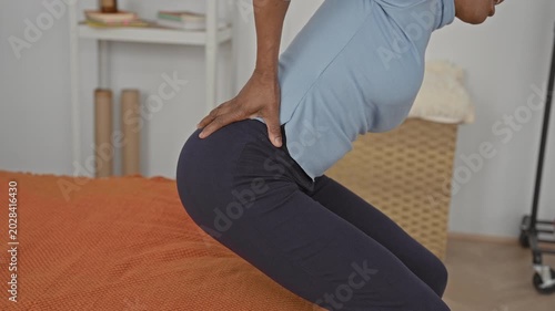 Woman sitting on bed holding lower back and leaning forward on orange bedcover in building, visible hip and hand contact with back; pain recovery.