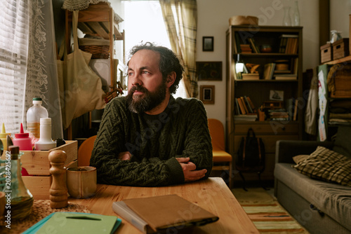 Obraz na plátně Middle aged Caucasian man sitting at table, looking through window, reflecting on financial hardship and uncertainty
