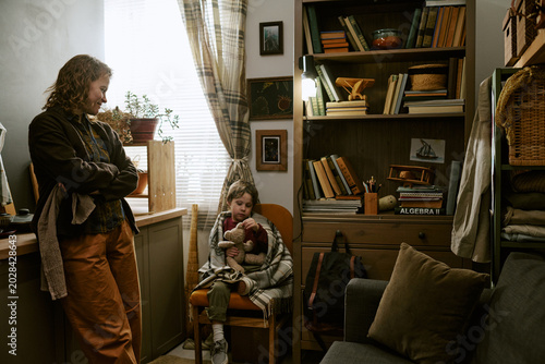 Konstfotografi Middle aged Caucasian woman standing with child sitting wrapped in blanket and holding teddy bear, showing poverty and hardship