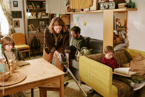 Middle aged Caucasian woman vacuuming living room while family reading and working at home, showing low income household routine. Useful for poverty, budgeting, family finance themes