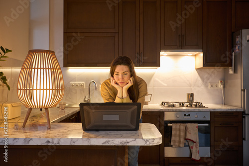 Woman working on laptop in kitchen at night with warm lamp light