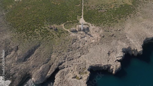 Aerial view of the white Plemmirio lighthouse standing on rugged rocky cliffs surrounded by green vegetation and a dirt path in Plemmirio, Sicily, Italy.