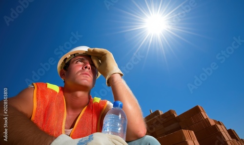 Young construction worker takes a moment to cool down, looking up at the bright sun, with sweat on his brow and a water bottle in hand