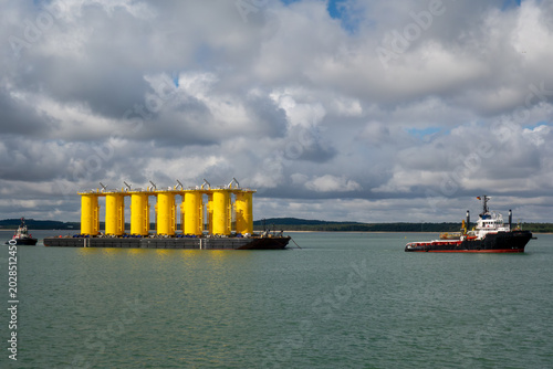 Offshore Wind Turbine Yellow Monopile Foundations on Transport Barge Towed by Tugboat at Sea. Renewable Energy Infrastructure Installation Project