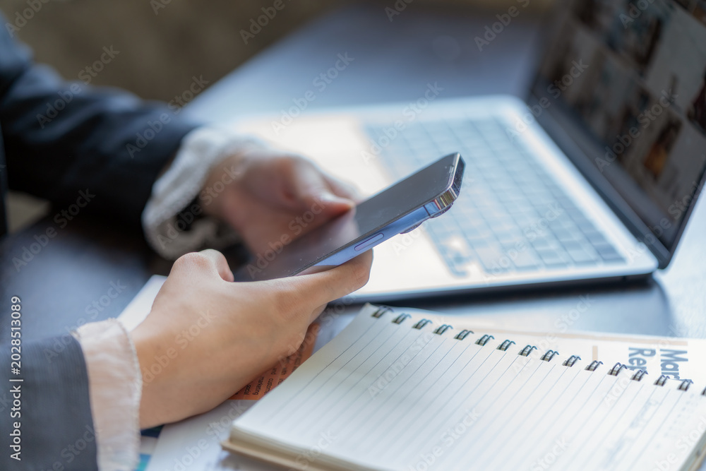 Fototapeta premium Close-up of a businesswoman using a smartphone with a laptop and notebook at an office desk.