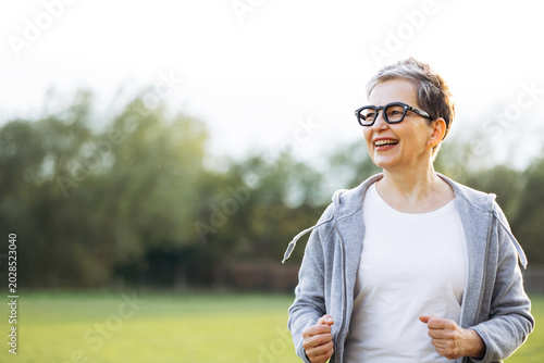 Active senior woman with grey hair wearing eyeglasses and sportswear, smiling while exercising outdoors in a park