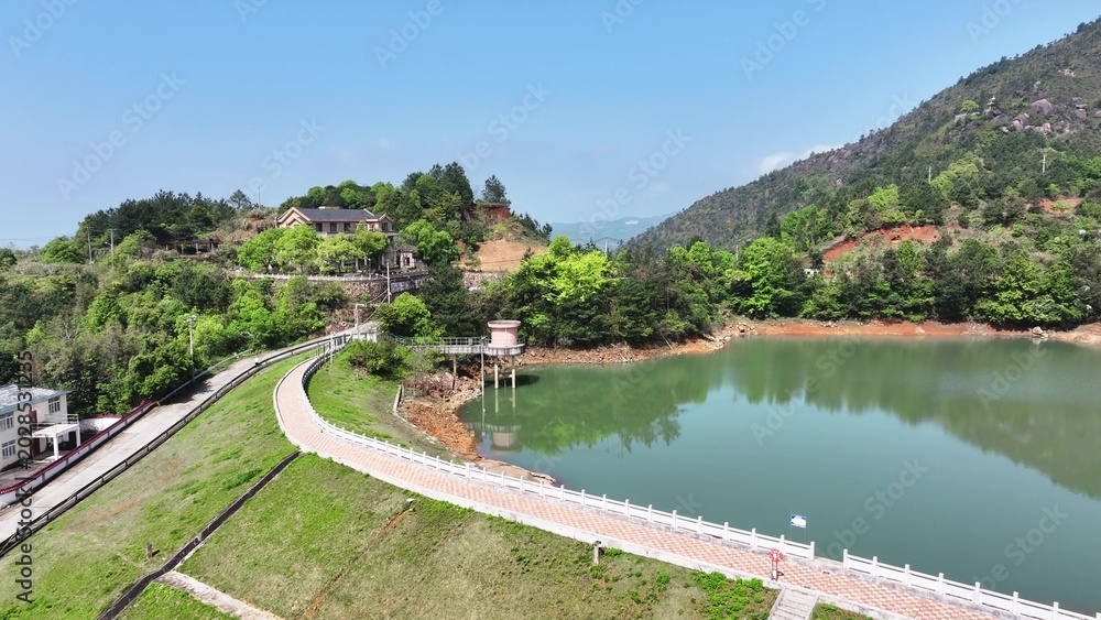 Fototapeta premium Winding dam and green lake surrounded by lush mountains under blue sky