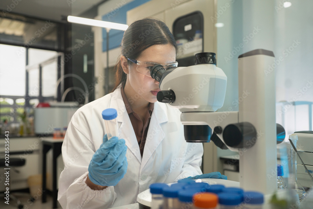 Fototapeta premium Female Scientist Looking Through Microscope While Holding Test Tube in Modern Research Laboratory