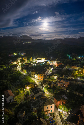 Aerial night view of Agioi Douloi village in Corfu, Greece, showing illuminated houses and streets nestled in the mountains under a dramatic cloudy sky and full moon.