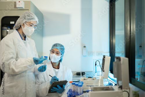 Tableau sur toile Two Asian female lab technicians in full PPE examine sample tubes and record dat