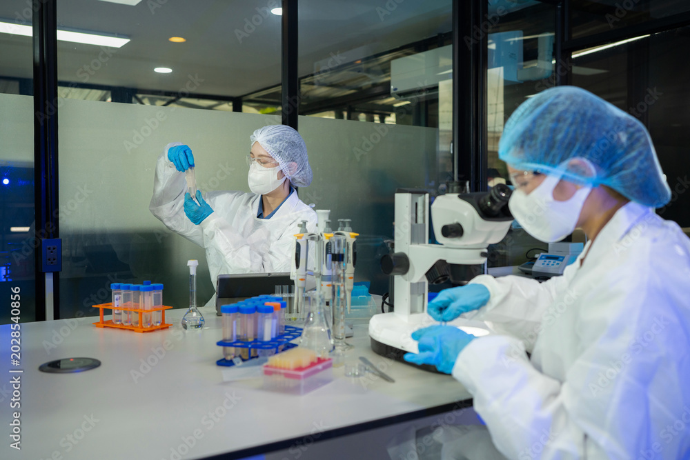 Naklejka premium Two lab technicians wearing white coats, masks, and gloves analyze specimens beside a microscope in a professional biomedical research facility