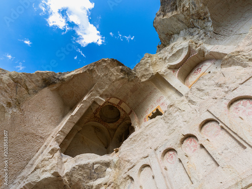 Exterior view of a historic rock-cut cave church carved into volcanic tuff in Göreme, Cappadocia, Türkiye. Ancient Christian architectural details, carved niches and faded red wall decoration traces a