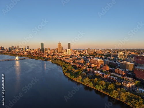 Golden hour aerial drone view of the Boston skyline, featuring the Prudential and John Hancock towers rising above the historic Back Bay neighborhood and the calm waters of the Charles River