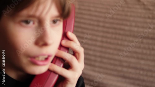 A close-up of a boy dialing a number on the keypad of a red retro telephone. The child brings the vintage receiver to his ear and enthusiastically begins to talk.