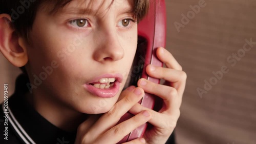 A close-up of a boy dialing a number on the keypad of a red retro telephone. The child brings the vintage receiver to his ear and starts a serious conversation.