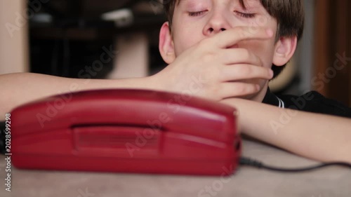 A bored boy yawns sleepily while waiting for a phone call, leaning his head on his hands in front of a landline device. A sad child waits for a message, looking at a red vintage retro telephone.
