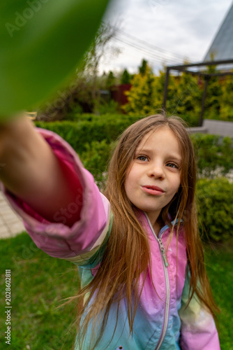 Engaging close-up portrait of thoughtful young girl in vibrant pastel jacket, capturing unique low-angle selfie outdoors in lush green garden with foreground leaf.