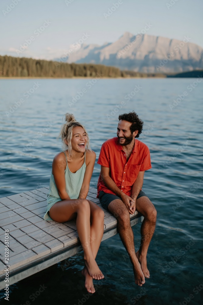 Fototapeta premium Couple sitting laughing on wooden dock by lake with mountain backdrop