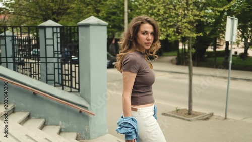 A stunning young woman looks back at the camera while walking down city steps, holding her blue shirt loose. Female portrait, urban lifestyle, casual attire, street view, confident gaze, tracking