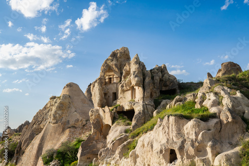 Historic rock-cut cave architecture carved into volcanic tuff formations in Göreme, Cappadocia, Türkiye. Ancient cave rooms and natural rock formations are surrounded by spring greenery under a blue s